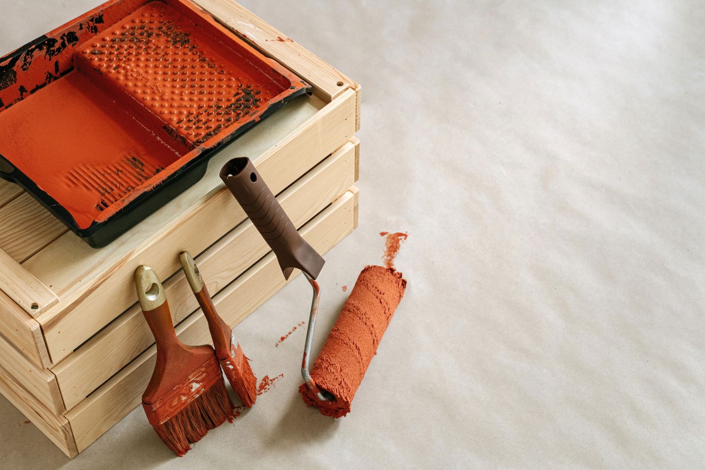 Bac, pinceaux et rouleau couverts de peinture orange près d’une caisse en bois.