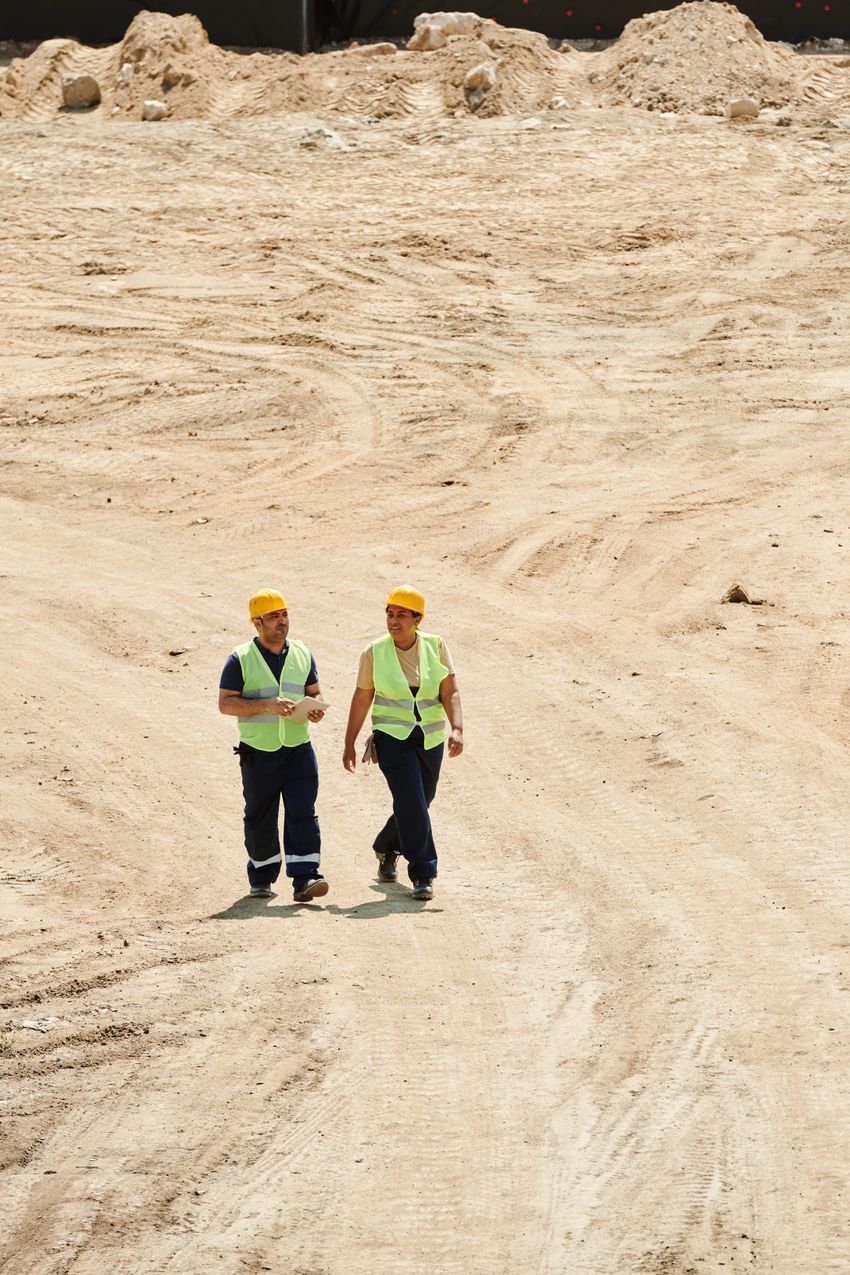 Two people wearing reflective vests and yellow hard hats walk across a construction site.