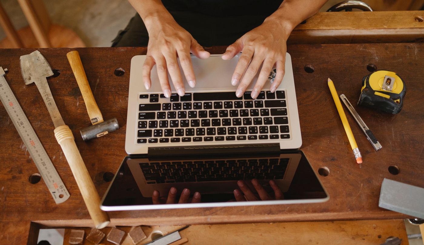 Dłonie na klawiaturze laptopa ustawionego na drewnianym stole warsztatowym wśród narzędzi stolarskich.