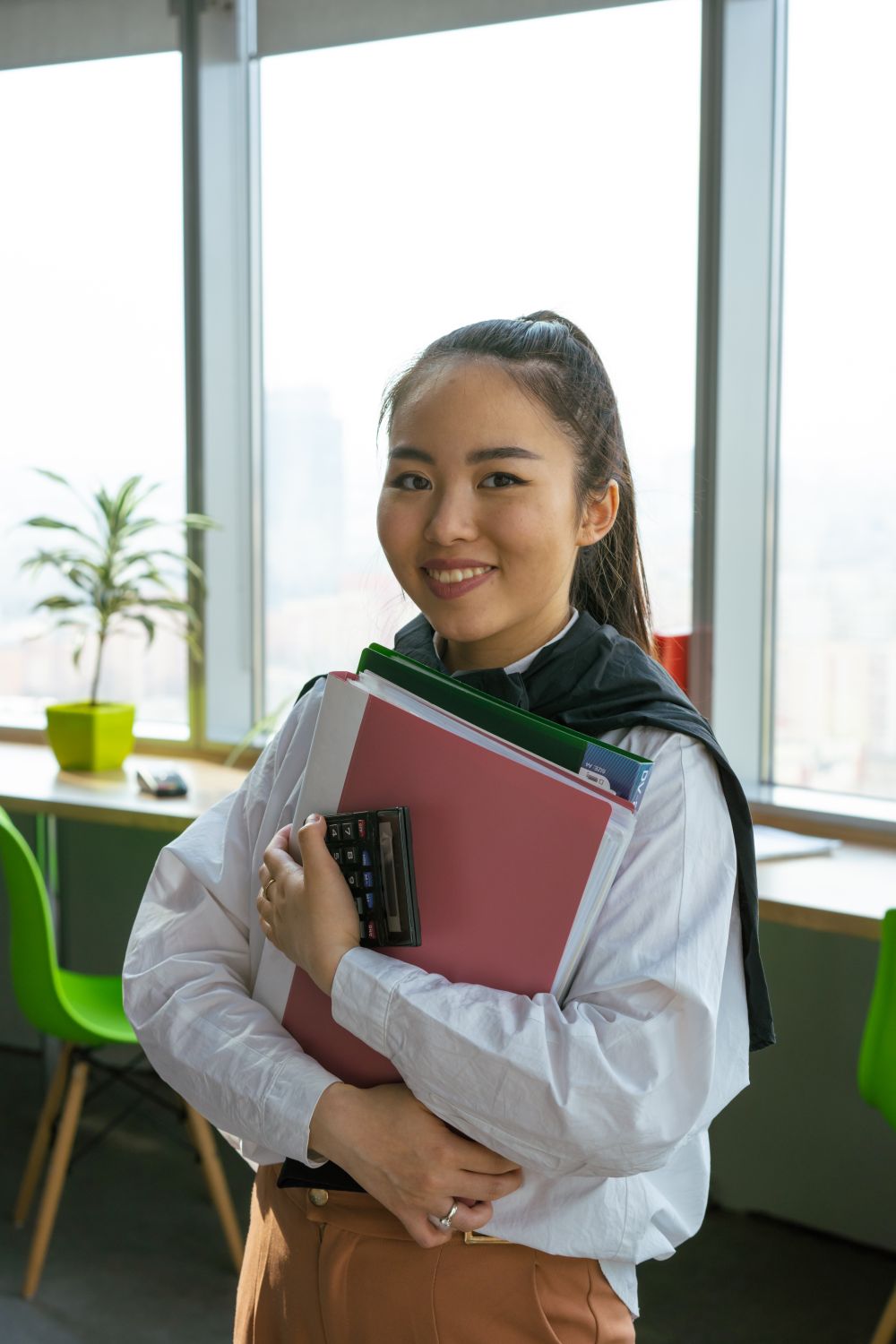 A woman holds folders and a calculator in an office.