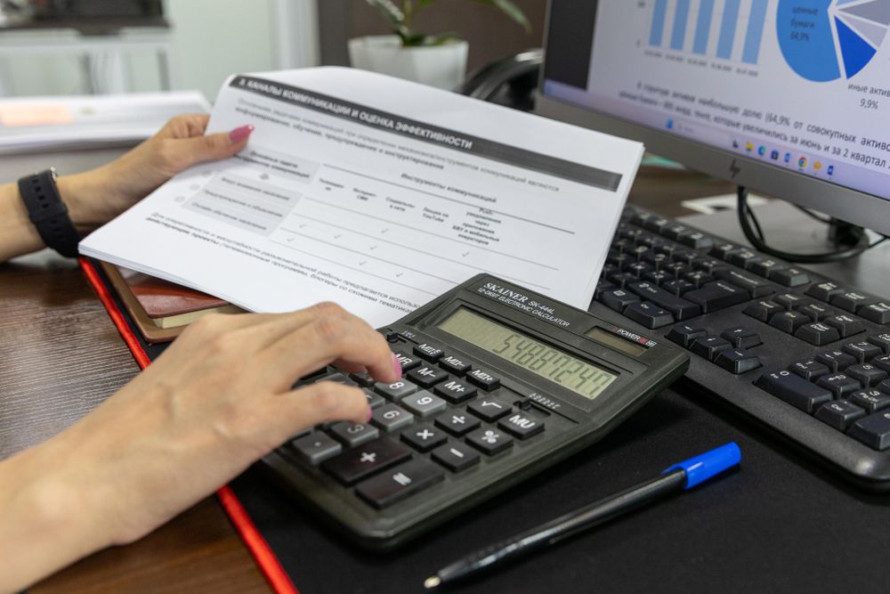 Mãos utilizam uma calculadora diante de documentos e de um ecrã.